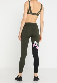 Zelené sportovní legíny ve stylu olivové barvy s těsným střihem, které mají černý panel a růžový grafický text na dolní části nohy. Hladký, pružný materiál.