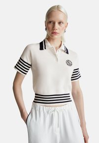 Mladá žena v bílém pleteném polo s černými pruhy na límci, rukávech a pase, v kombinaci s bílými kalhotami na stahování, stojící s rukou v kapse.