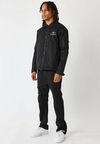 Sort jakke med foran lynlås, flere lommer og logo på brystet; parret med matchende bukser og hvide sneakers. Glat, slidstærkt stof.
