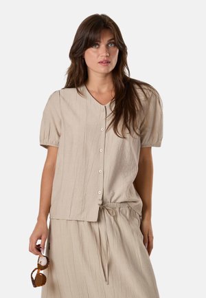 Femme aux longs cheveux bruns portant un chemisier beige à manches courtes boutonné et une jupe assortie, tenant des lunettes de soleil écaille dans une main.