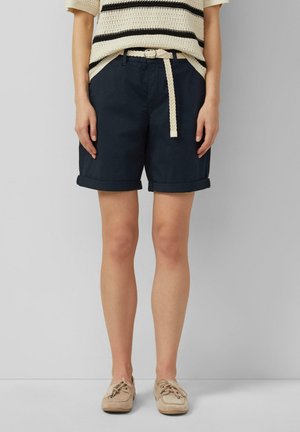 Persoon draagt marineblauwe korte broek met opgerolde zoom, een crème gevlochten riem, een crème en zwart gestreepte gebreide top en beige mocassins en staat.