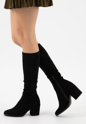 Jambes portant des bottes en daim noir montantes jusqu'aux genoux avec des talons carrés et des fermetures éclair latérales, associées à une jupe plissée vert olive.