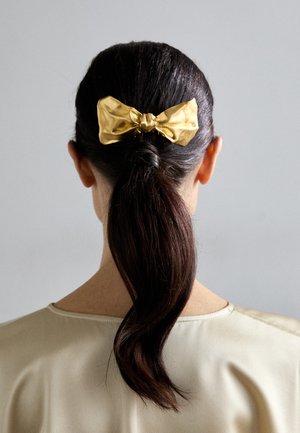 Χρυσό σατέν φιόγκο μαλλιών με κόμπο στη μέση, τοποθετημένος σε σκούρα, ίσια, μεσαίου μήκους μαλλιά, φορεμένος με ένα ανοιχτό μπεζ μπλουζάκι, απέναντι από έναν γκρι φόντο.