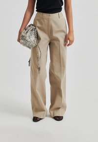 Pantaloni beige a vita alta con una vestibilità rilassata e pieghe, abbinati a una borsa a tracolla con stampa serpente testurizzata e décolleté in mesh nero con punta aperta.