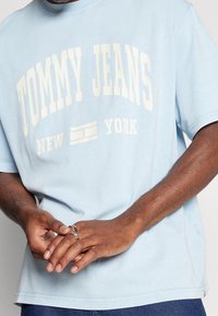 Camiseta de algodón azul claro con "TOMMY JEANS" y "NUEVA YORK" impresos en color crema. Mangas cortas, corte relajado, tejido texturizado y costuras laterales visibles.