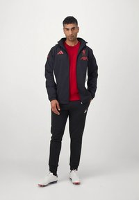 Juoda sportinė striukė su raudonomis detalėmis, trumpu apykakle ir priekinio užtrauktuko uždarymu. Po ja – raudonas džemperis, juodos sportinės kelnės. Baltas sportinis bateliai.