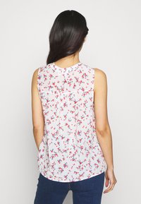 Blusa sin mangas blanca y transparente con un patrón floral en rojo y azul, con la parte trasera plisada y dobladillo redondeado, combinada con jeans oscuros.