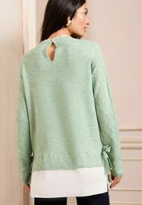 Mintgrüner Pullover mit Spitzenapplikationen an den Ärmeln, einem Schlüsselloch-Ausschnitt hinten und gebundenen Akzenten am Saum, übereinander getragen mit einem weißen Hemd.