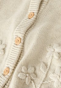 Kreemjas kootud kardigan soonikkoega krae, puidust nööpide ja lillelise tikandi detailidega. Pehme tekstuur ja soe visuaalne mulje.