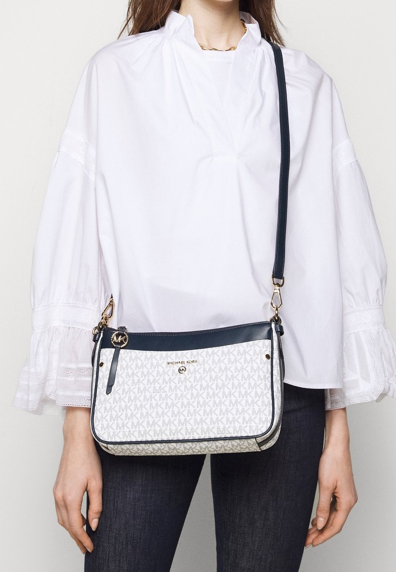 Femme portant un chemisier blanc et un jean foncé porte un sac bandoulière Michael Kors blanc et bleu marine avec motif logo et accents dorés.
