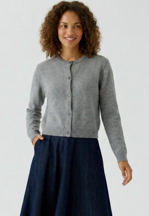 Vrouw met krullend haar die glimlacht, een lichtgrijze vest met knopen en een donkerblauwe rok draagt, staand met één hand in de zak.