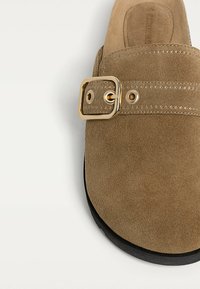 Brown suede slip-on shoe with a wide strap, featuring a rectangular gold buckle and stitched accents, complemented by a black sole.