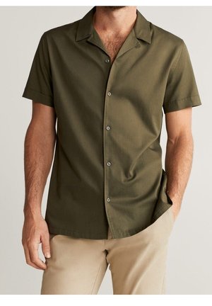 Homme portant une chemise à manches courtes boutonnée col olive et un pantalon beige, une main dans la poche, sur un fond neutre.