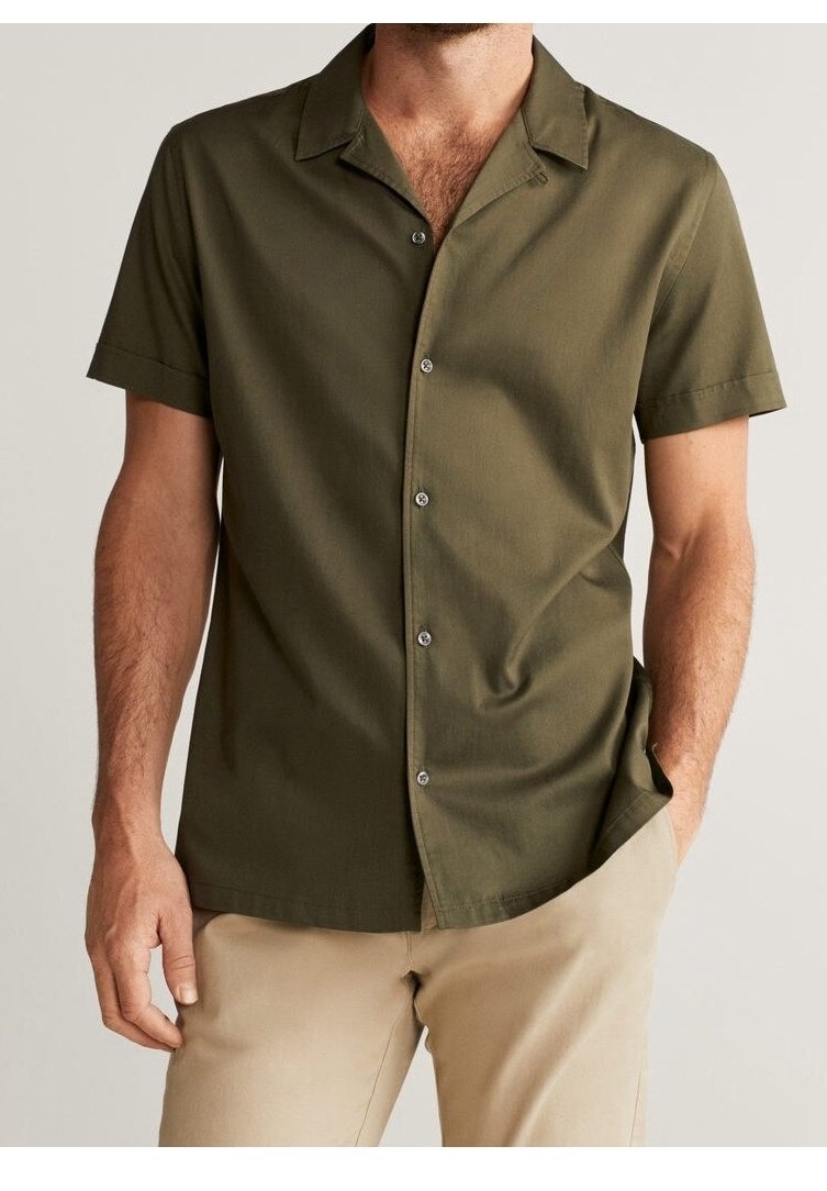 Homme portant une chemise à manches courtes boutonnée col olive et un pantalon beige, une main dans la poche, sur un fond neutre.