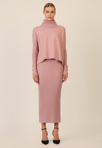 Rosa gerippter Strickkleid mit einem halben Stehkragen und einem lässigen, cropped Pullover. Mit Knopfakzenten an den Ärmeln versehen, kombiniert mit schwarzen High Heels.