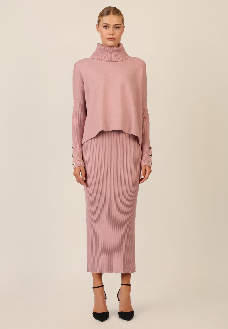 Rosa gerippter Strickkleid mit einem halben Stehkragen und einem lässigen, cropped Pullover. Mit Knopfakzenten an den Ärmeln versehen, kombiniert mit schwarzen High Heels.
