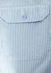 Tessuto a righe blu chiaro con sottili linee bianche, con una tasca sul petto e un bottone blu in cima. La superficie texturizzata è liscia.