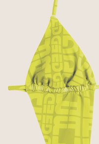 Keltainen bikinitoppi, jossa on kolmion muotoinen muotoilu, keskellä poimutus ja toistuva logokuvio. Ohut olkaimet, jotka tarjoavat säädettävän istuvuuden.