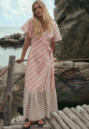 Lyshåret kvinde i stribet omslagskjole og sandaler læner sig på tovgelænder ved stenede kyst med hav og overskyet himmel i baggrunden.