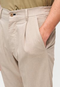 Pantalon beige texturé avec plis, fermeture à bouton et poches latérales. Le matériau semble léger et respirant.
