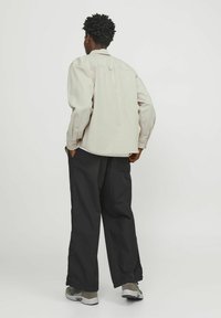 Chemise oversized beige clair avec un col de chemise, des poignets boutonnés et un empiècement au dos, associée à un pantalon noir ample et des baskets grises.