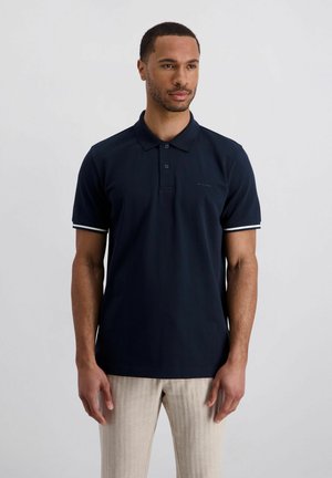 Mann steht, trägt ein dunkelblaues Poloshirt mit weißem Besatz an den Ärmeln und hellbeige Hosen, vor einem schlichten hellen Hintergrund.