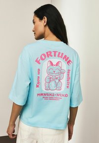 Svetlo modra prevelika majica s kratkimi rokavi z grafičnim motivom valovajoče mačke v roza in beli barvi na hrbtni strani, z napisi "FORTUNE" in "prinaša srečo".