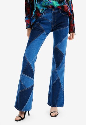 Femme portant un jean évasé en patchwork de différentes teintes de denim, associé à des sandales à talons hauts noires à lanières et un haut coloré à motifs.