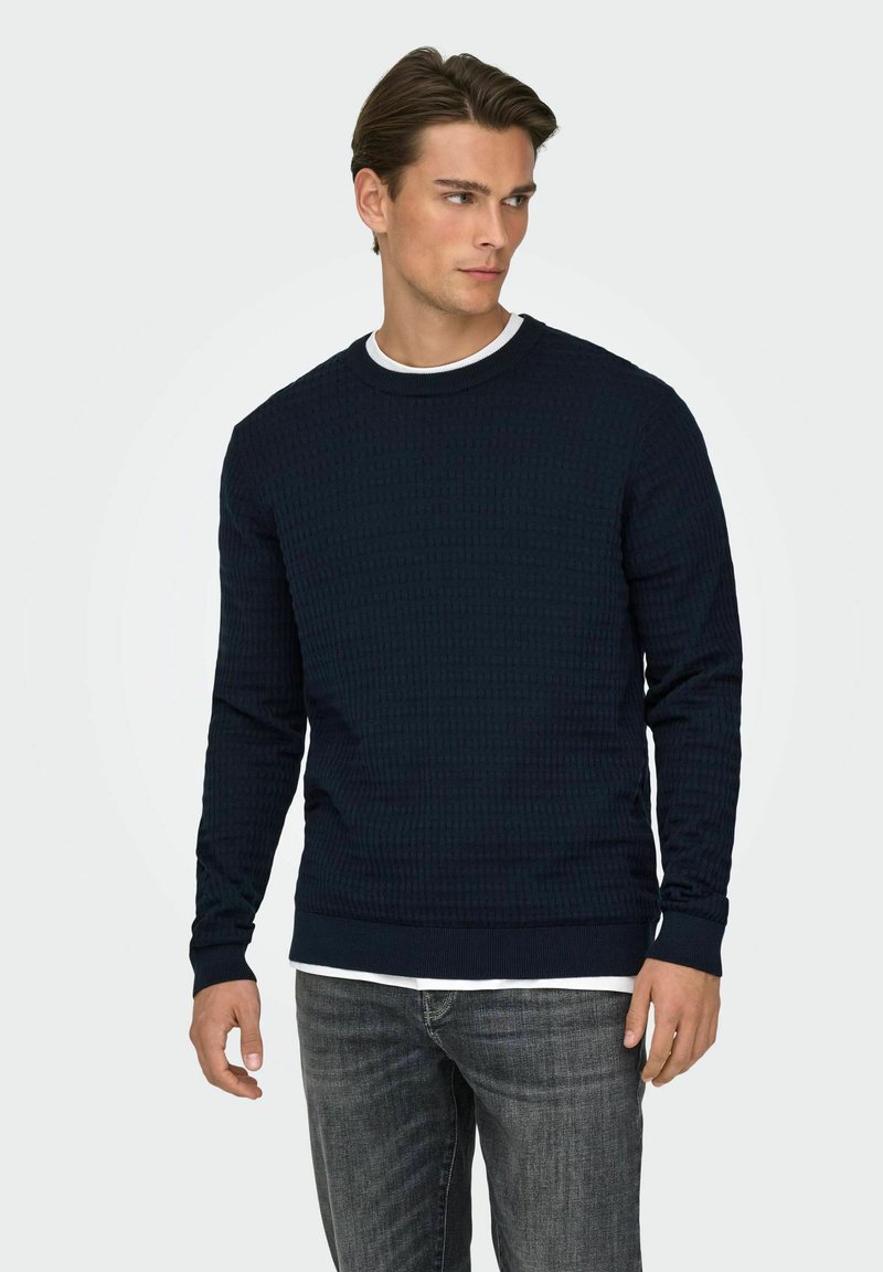 Sweat-shirt bleu marine texturé à manches longues avec poignets et encolure côtelés, superposé à une chemise blanche. Porté avec un jean gris droit.