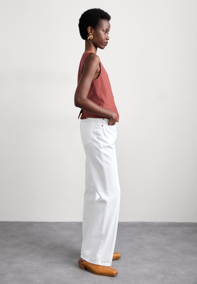 Top sin mangas de color óxido con un diseño estructurado, combinado con jeans blancos de pierna ancha y zuecos de tacón marrones. Estética simple y moderna.