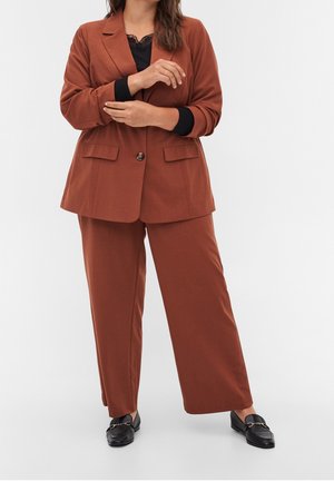 Femme portant un costume couleur rouille avec pantalon à jambes larges, haut noir bordé de dentelle et mocassins noirs, debout les bras croisés.