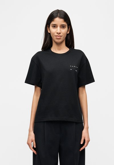 Μαύρο βαμβακερό t-shirt με άνετη εφαρμογή, στρογγυλή λαιμόκοψη, κοντά μανίκια και λευκό γραφικό τύπωμα στο στήθος. Φορεμένο με σκούρο παντελόνι με λεπτές ρίγες.