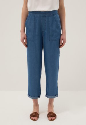 Donna che indossa pantaloni corti blu con tasche anteriori e risvolti arrotolati, abbinati a sandali marroni intrecciati.