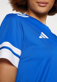 Camisola atlética azul feita de tecido suave, com riscas brancas nas mangas e um logotipo branco no peito.