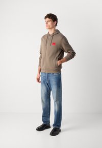 Sudadera marrón con cremallera y un parche de marca rojo, combinada con unos vaqueros de color azul claro y zapatos negros. El modelo se encuentra de pie frente a un fondo liso.
