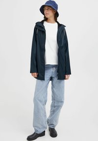 Navy waterproof rain jacket with adjustable hood, paired with a white t-shirt, light blue jeans, and black ankle boots, worn with a blue bucket hat.