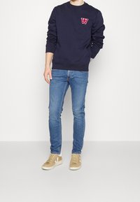 Marinblå sweatshirt med rött bokstavslogo, blå slim-fit jeans och beige sneakers med vita detaljer. Tyget ser mjukt ut; avslappnad passform överlag.