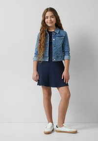 Jeansjacke mit Knöpfen, blaue Farbe; navyblaues gestreiftes Kleid; weiße und navyblaue Sneakers mit beiger Sohle. Junge weibliche Model posiert.