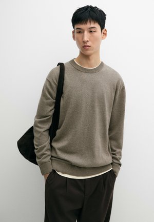 Giovane uomo con capelli corti neri, che indossa un maglione beige, pantaloni scuri e una borsa a tracolla nera, in piedi davanti a uno sfondo bianco uniforme.