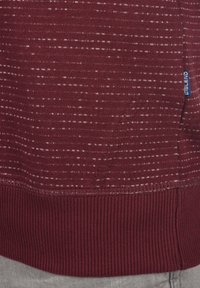 Burgunderfarbener Strickpullover mit einem strukturierten Muster aus weißen Streifen, mit einem gerippten Saum und einem kleinen blauen Etikett an der Seite.
