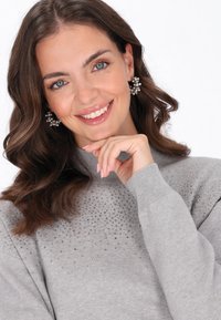 Pulover gri cu gât înalt, cu strasuri împrăștiate pe umeri. Purătoarea are părul ondulat și își atinge bărbia.