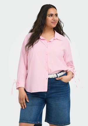 Donna con lunghi capelli scuri che indossa una camicia rosa chiaro abbottonata con polsini annodati e pantaloncini di jeans blu fino al ginocchio con cintura di pizzo bianca.