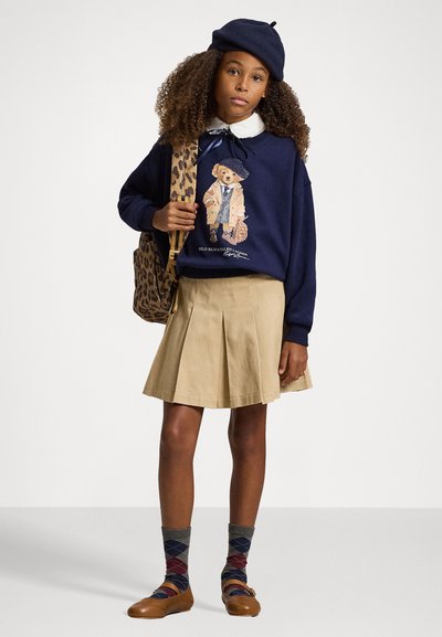 Sudadera navy con un gráfico de oso, falda con pliegues color beige, mochila de estampado animal y calcetines de rombos con zapatos marrones.