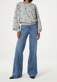 Osoba oblečená v šedé mikině s modrými srdíčky, v širokých modrých džínách a černých botách s špičatou špičkou na jednolitém pozadí.