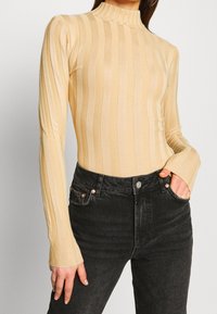 Top a maniche lunghe beige a coste con collo alto, caratterizzato da strisce verticali e maniche leggermente svasate, abbinato a jeans di denim scuro.