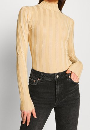 Beige ribbet trøje med lange ærmer og en mock neck, der har lodrette striber og let udvidede manchetter, parret med mørke denimjeans.
