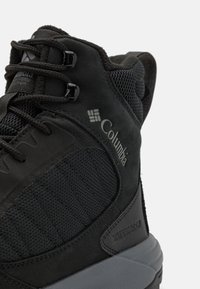 Bota de senderismo negra impermeable con parte superior de malla texturizada, puntera reforzada y un sistema de cordones seguro con un logo distintivo en el costado.