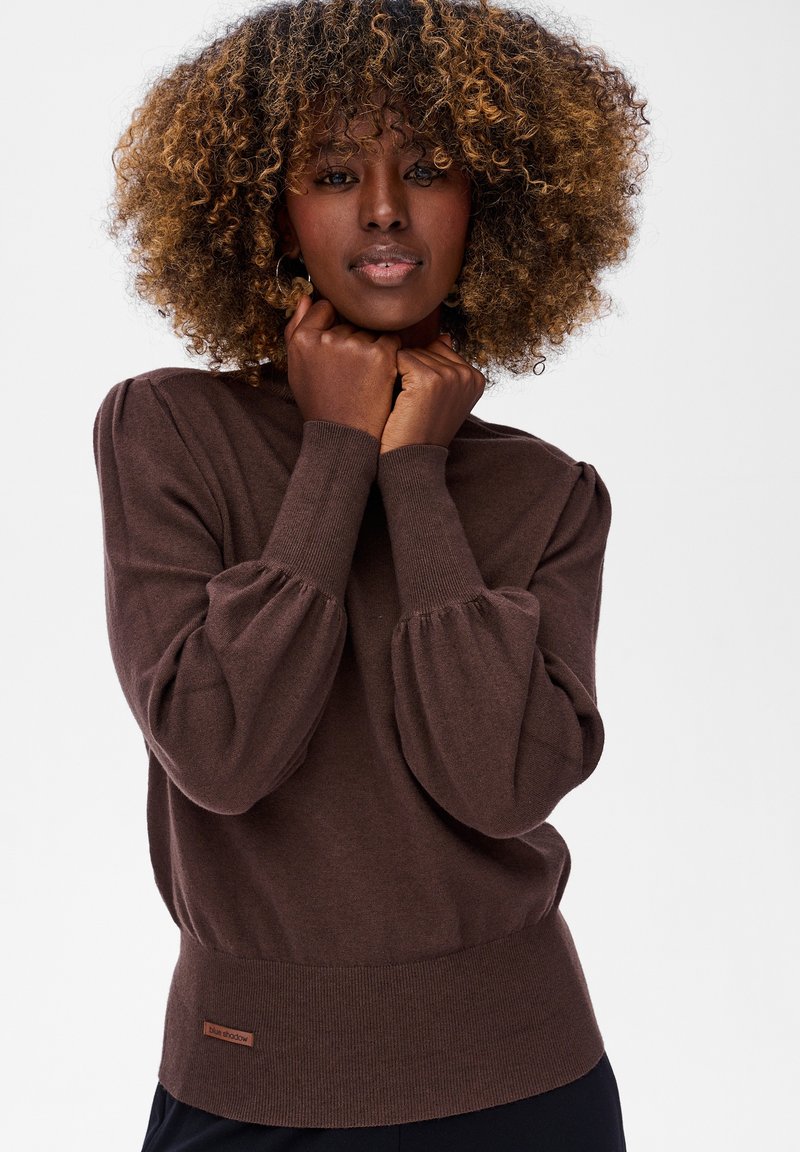 Kobieta z kręconymi włosami, ubraną w brązowy sweter z długim rękawem, trzymająca dłonie blisko brody, stoi na tle gładkiej białej ściany.