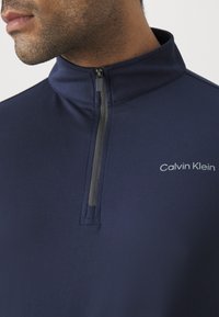 Marineblå hettegenser med glidelås laget av glatt og lett stoff. Har høy krage og grå logo på venstre bryst. Glidelås med svart trekk.