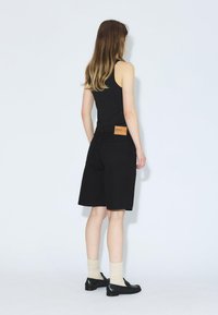 Bermudas de mezclilla negra con una etiqueta de cuero marrón, combinadas con una camiseta sin mangas negra, calcetines crema y zapatos negros, vistas desde atrás.